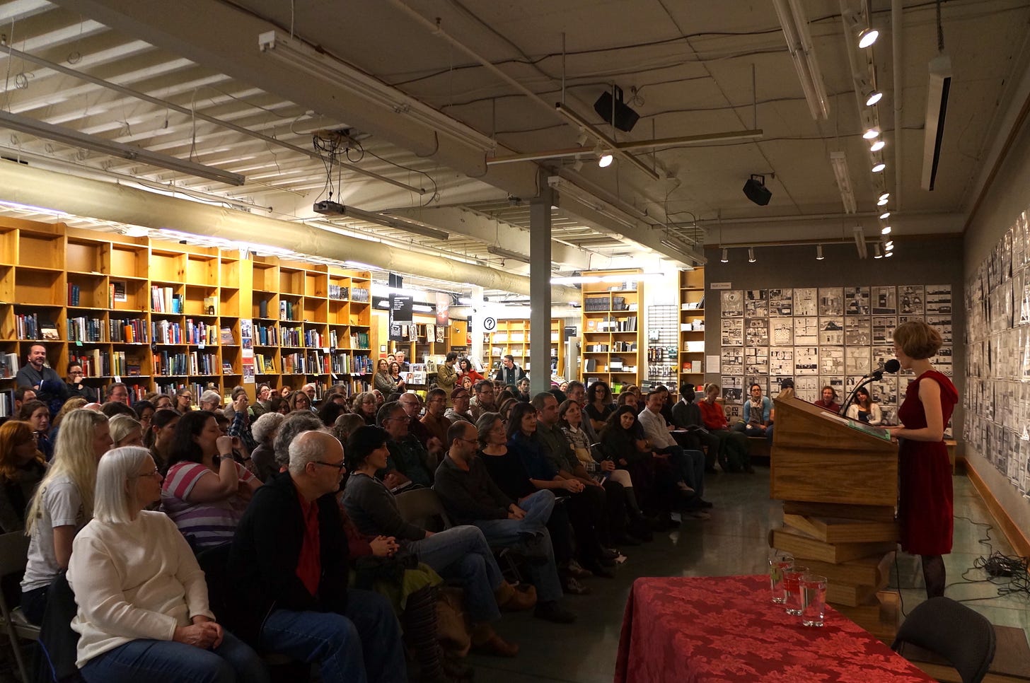 Woman with short hair and a red velvet vintage dress stands at the lectern at Powell's, with a crowd of people sitting in the audience. On the walls, Nicole J. Georges's art in black and white tells another story. 