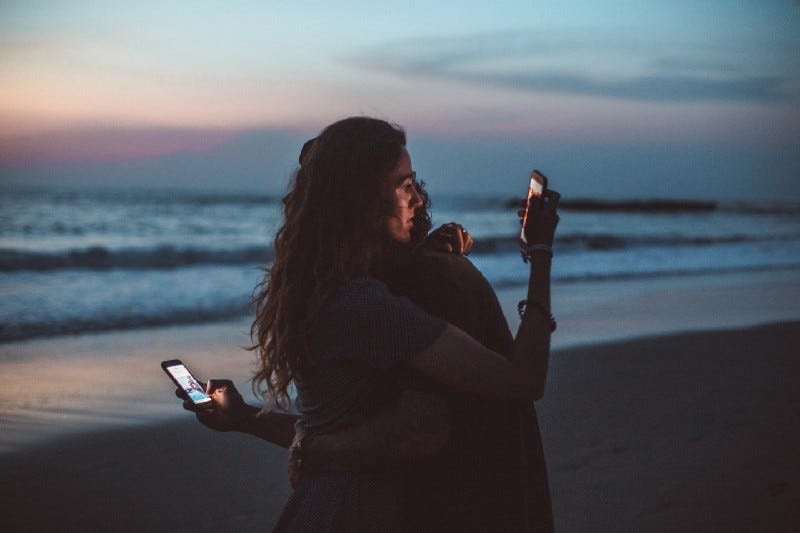 Two people hug on a beach while looking at their phones.