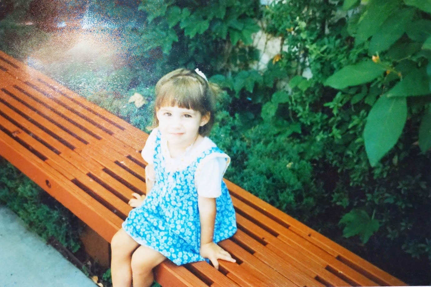 A very tiny Isa, around 5 or 6 years old, sits in her grandmother's backyard. She's wearing a blue dress.