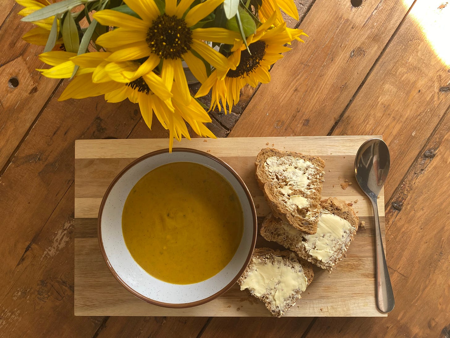 A bowl of soup on a wooden board, bread and butter to the side next to a spoon. Some yellow sunflowers are also on the table. 
