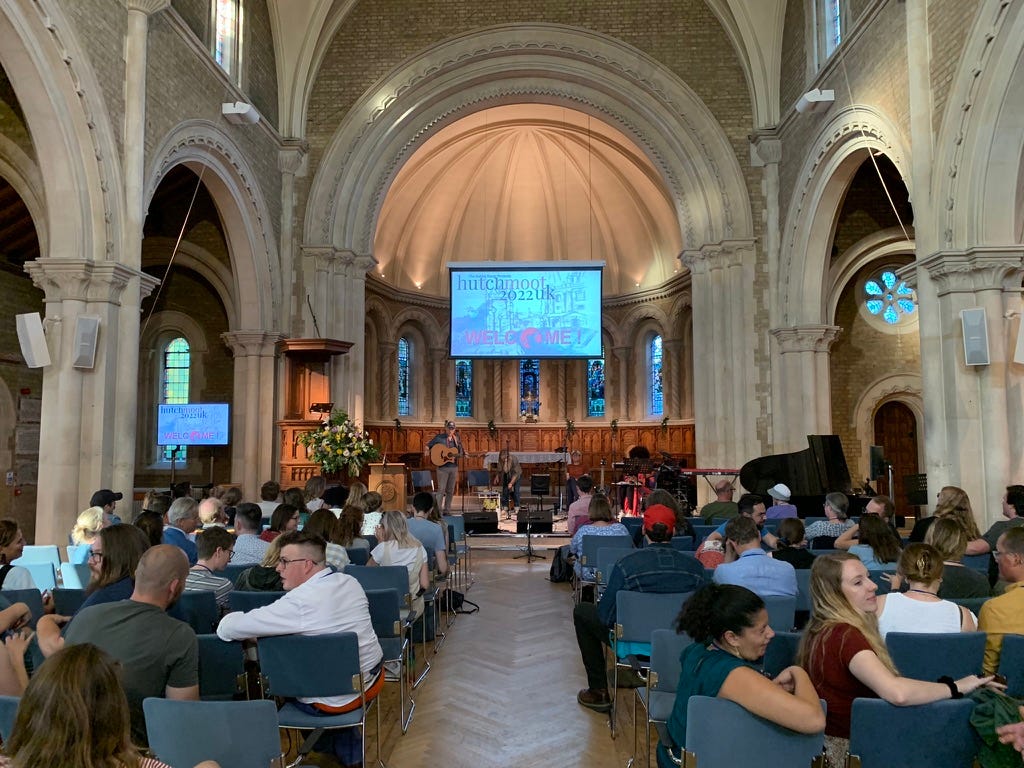 People sitting chatting in St Andrews Church while Andrew Peterson prepares his guitar, beneath the stone arch of the church, while a projector shows 'Hutchmoot 2022 UK - Welcome!'