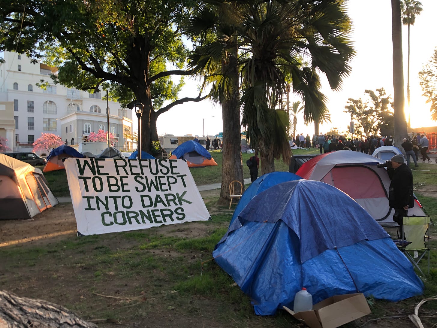 Tents in the park, and a sign that reads "WE REFUSE TO BE SWEPT INTO DARK CORNERS"