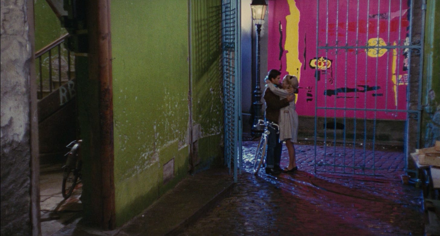 Geneviéve and guy sharing a tender moment in the alley outside of his apartment, with a bright pink and yellow wall in the background and a slightly less bright but still bright green apartment wall to their left