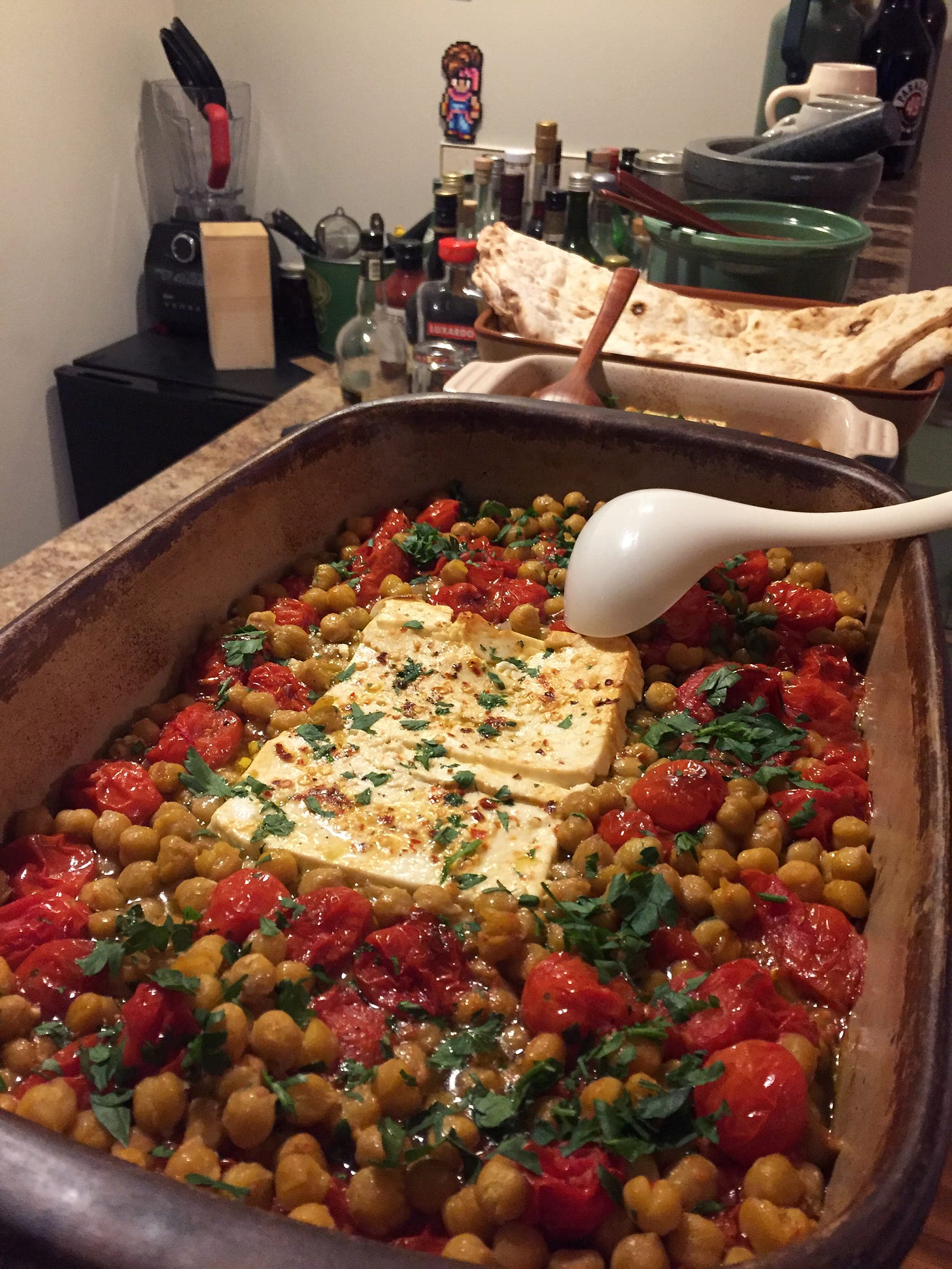 A large stoneware dish filled with the baked chickpeas, feta, and tomatoes, sprinkled with herbs and chili flakes. Behind the dish is a smaller baking dish with the tofu version, a plate of taftoon, and a green ceramic bowl containing the salad.