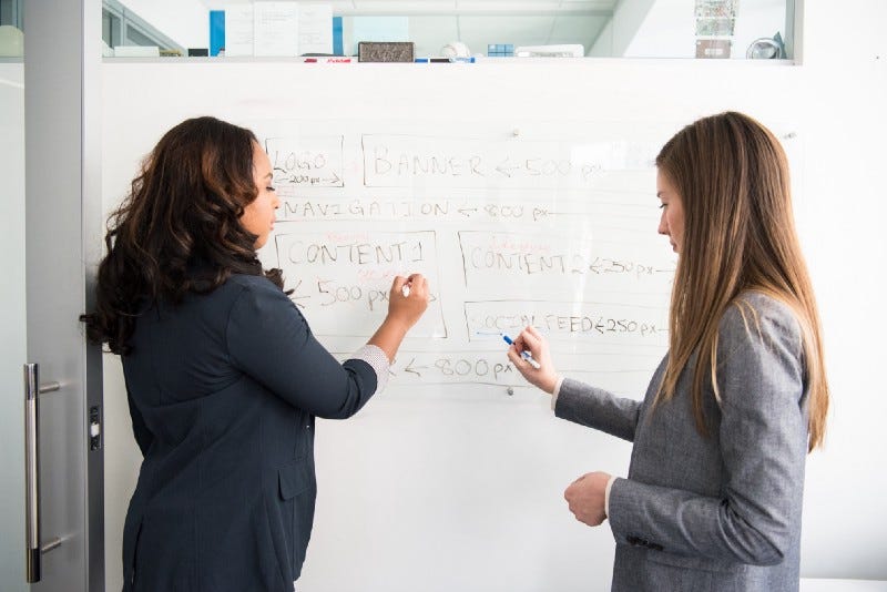 Two women talking in front of a white board