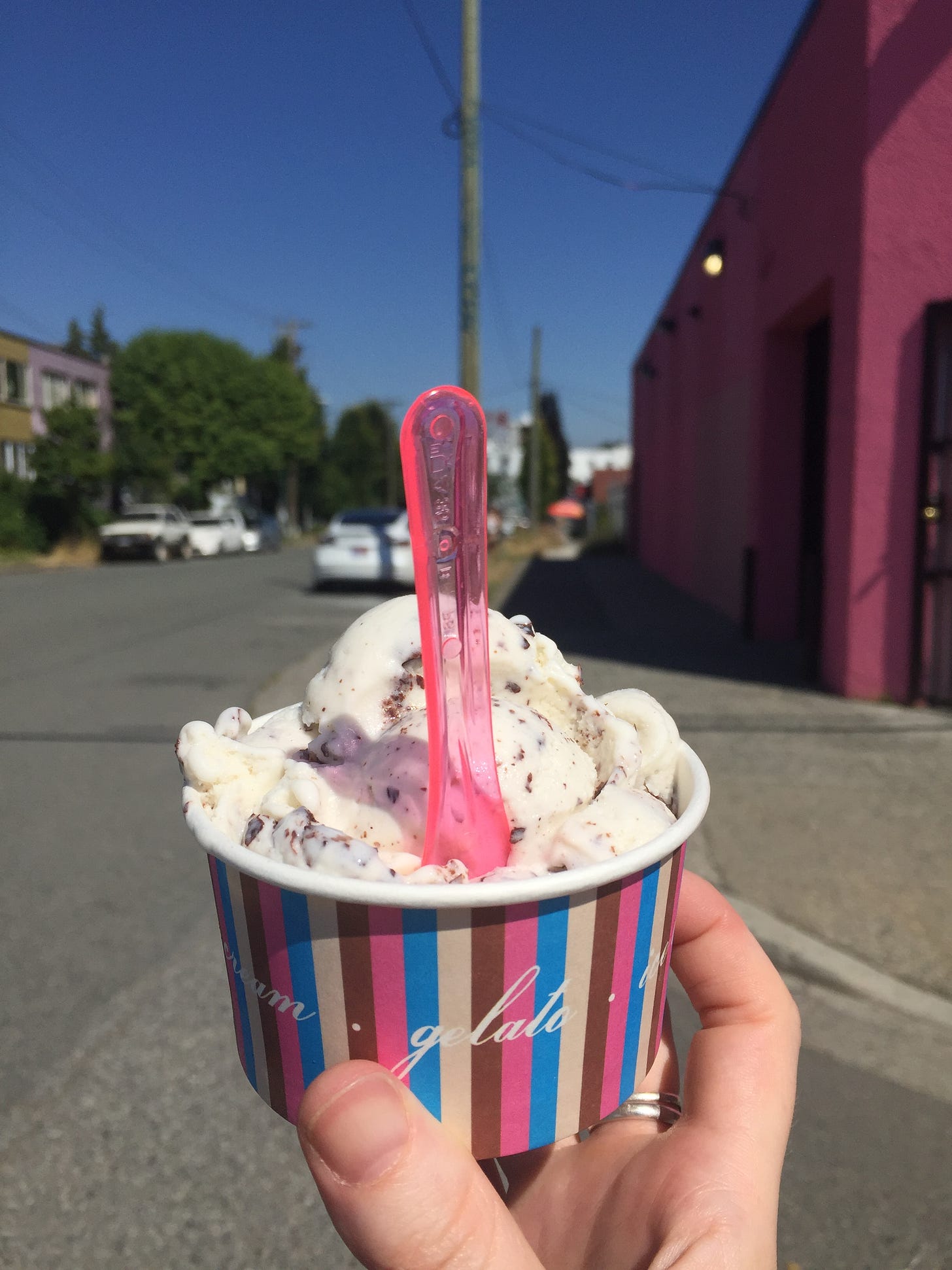 My hand holding a striped paper cup of stracciatella gelato with a clear pink spoon sticking out of it. In the background is a quiet street lined with a few parked cars, and a hot pink building is on the right hand side of the street. The sky is bright blue.
