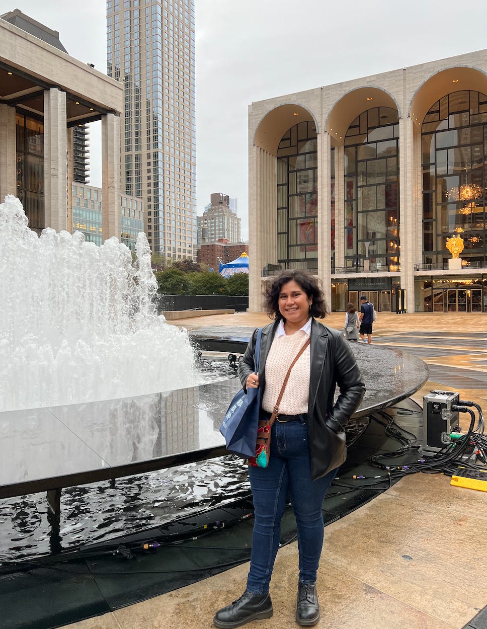 Alicia stands by the fountain in front of the Met Opera in New York City. Alicia has short dark curly hair and medium-brown skin. Alicia wears a couple of bags, and the clothing is a cream sweater over a white button-down, with a leather jacket and jeans and black leather boots. Alicia smiles. 