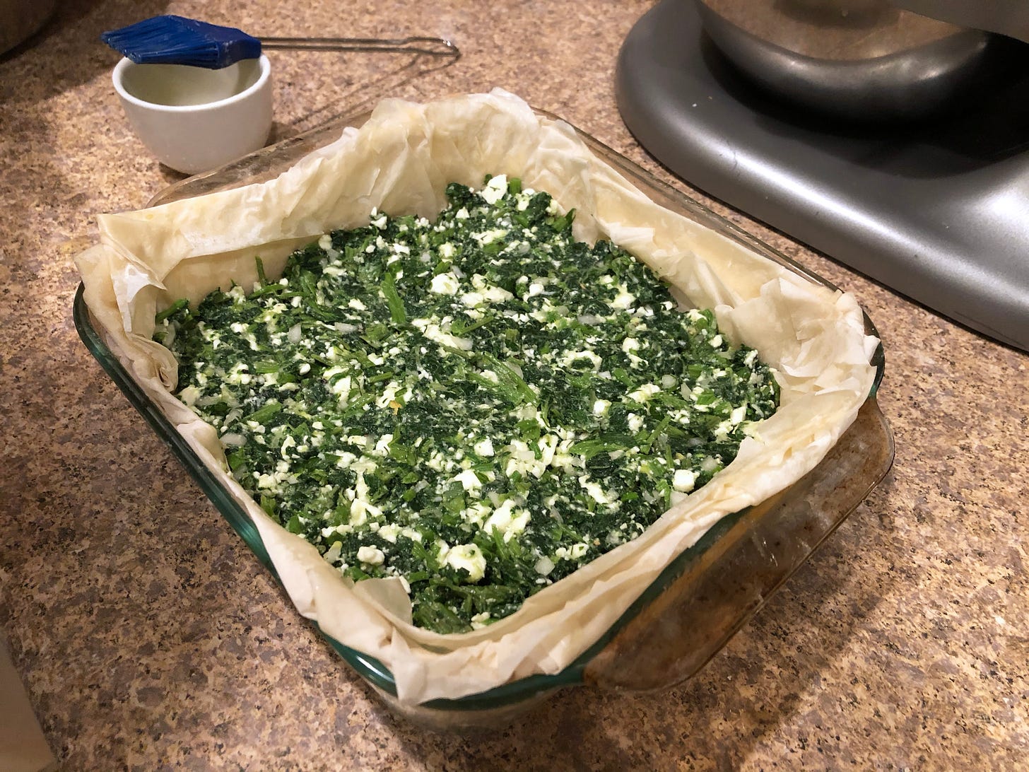 a square glass baking dish filled with sheets of pastry, which has green spanakopita filling pressed into it. a basting brush sits to the left of the dish.