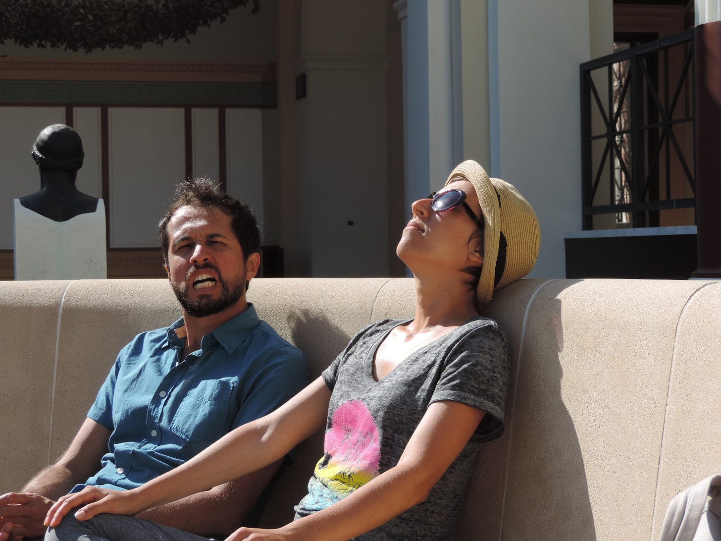 foto mostrando um cara e uma mulher sentados em um banco circular de concreto, ao sol, com ele olhando para a câmera e fazendo uma careta, enquanto ela toma sol com a cabeça reclinada para trás