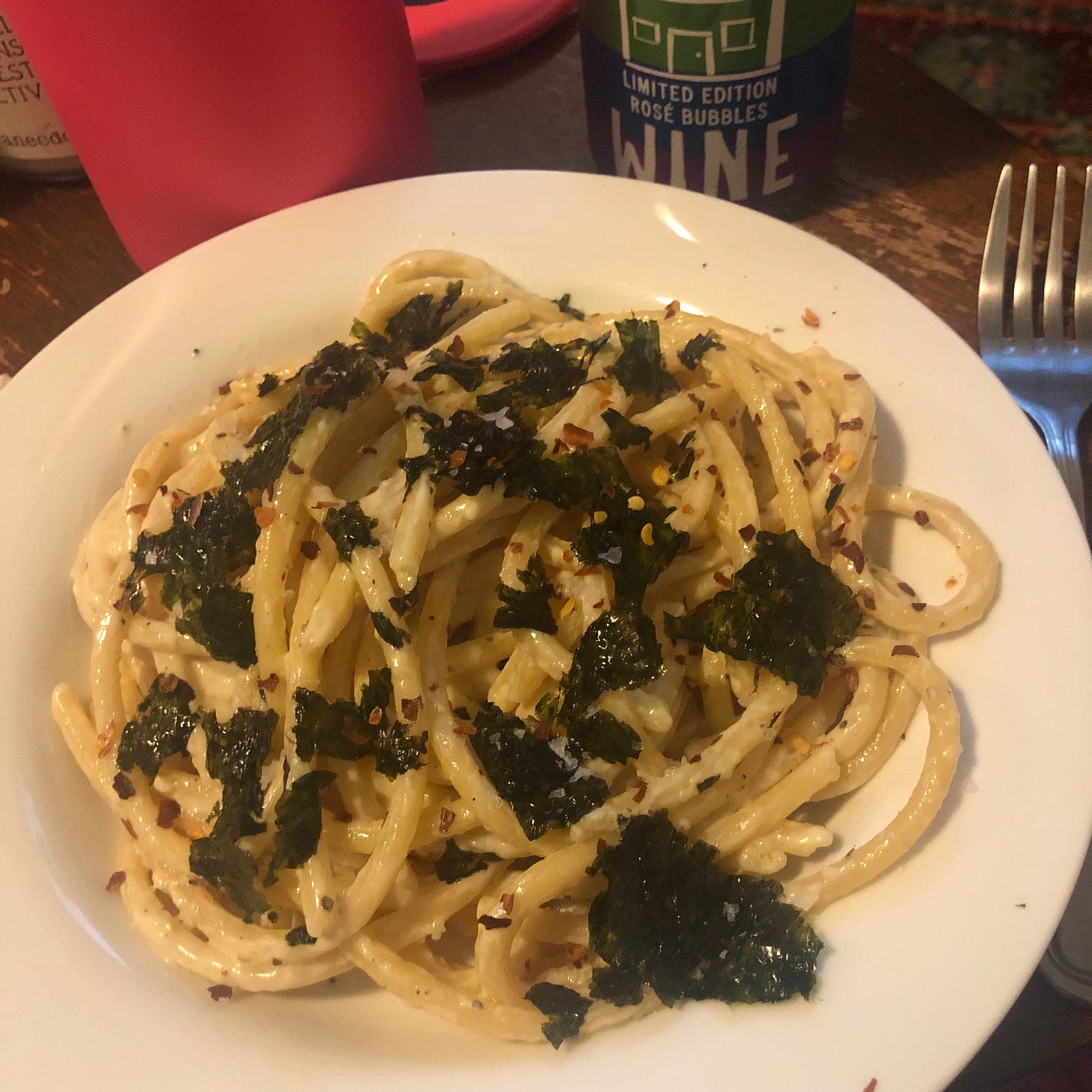Image: bucatini in cream sauce with roasted seaweed on top, sprinkled with red chili flakes and served with a canned sparkling rosé