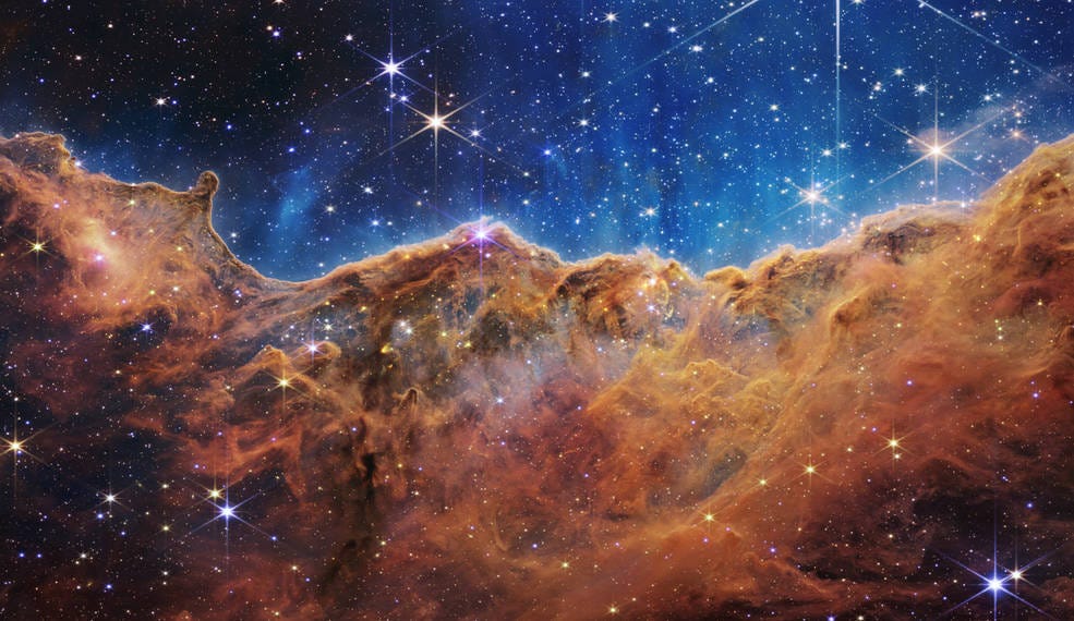 Photo of the Carina Nebula in space, showing a brownish cloud of gas against a dark blue space sky, the top of the photo studded with bright stars.