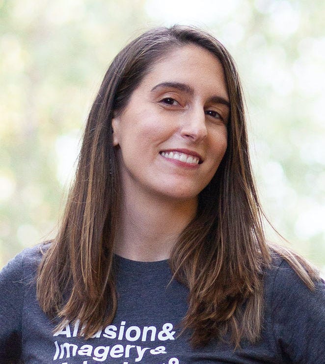 A white presenting woman with long, straight brown hair and brown eyes smiles into the camera while head is slightly facing to the right. She is wearing a dark gray t-shirt with the words Allusion& Imagery& showing in the frame.