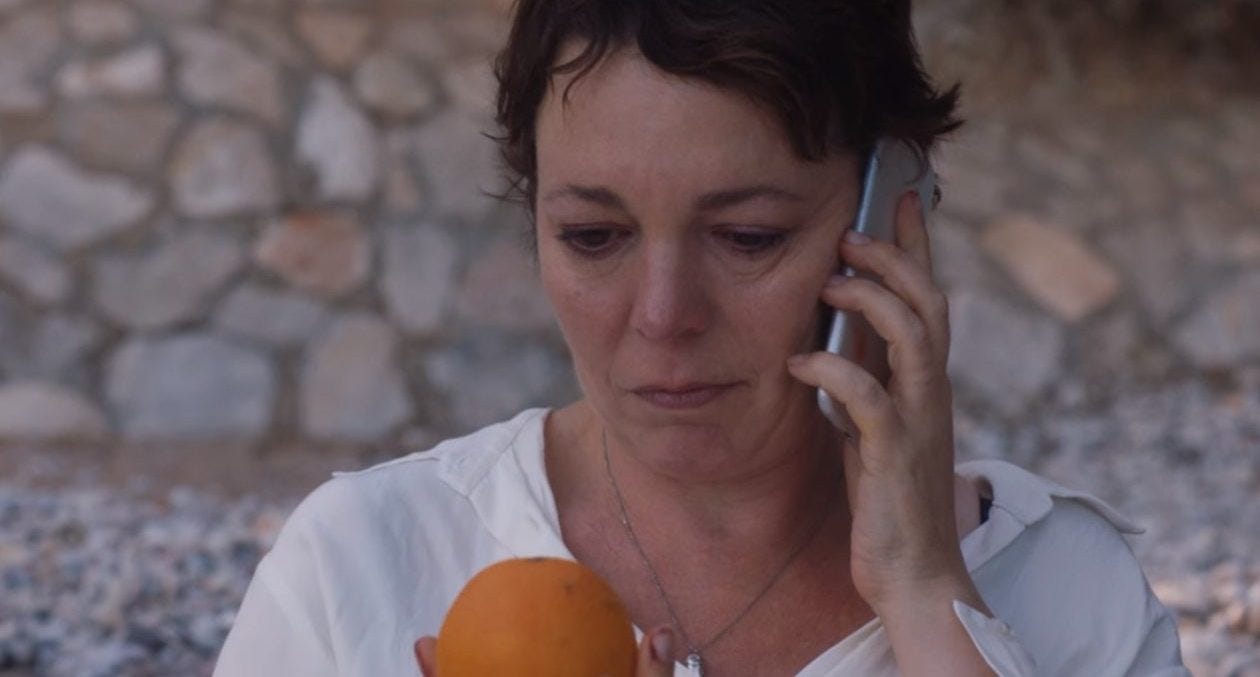 A still from 'The Lost Daughter' - A white middle aged woman holds a phone to her ear, while observing an orange held in her other hand. Her expression is inscrutable. 