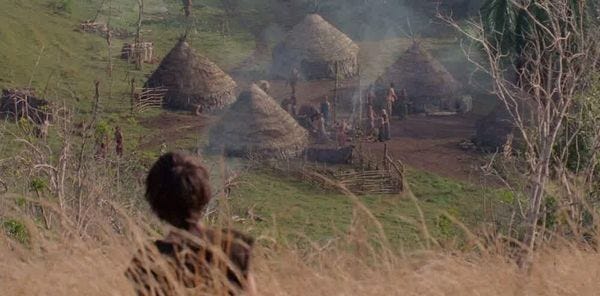 Brother surveils the human village, made of huts with thatched roofs.