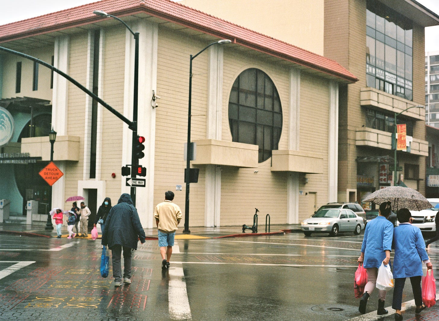 A multiple cross-walk in Oakland's Chinatown with different individuals and groups crossing. It's a rainy day.