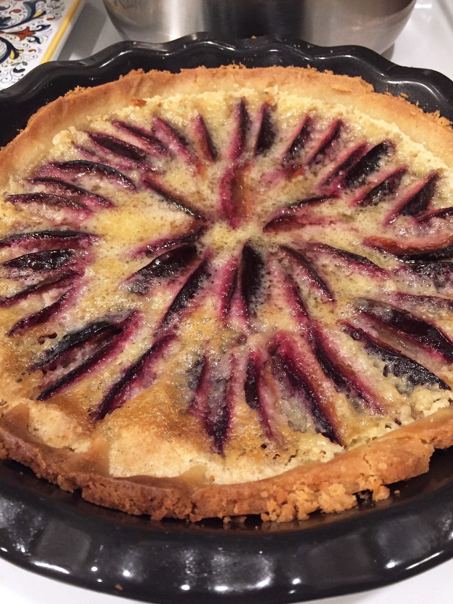 In a black ceramic pie pan, concentric circles of dark-skinned plum slices sit sunken into a frangipane tart with browned edges.