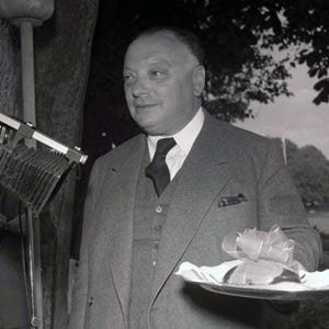 A black and white photo of a plump, middle aged physicist carrying a plate with some kind of ribbon on it.