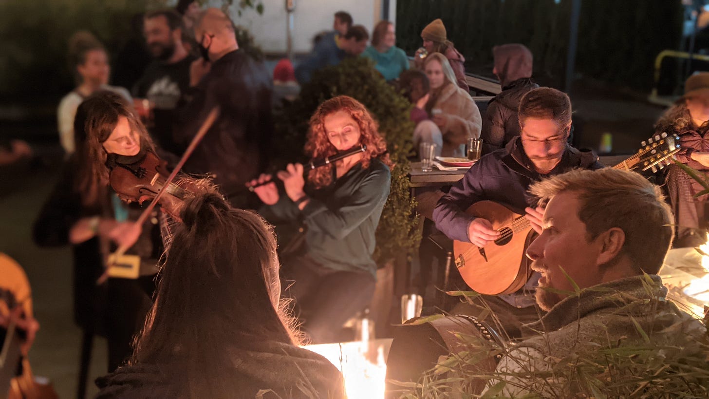 A fiddler, a flute player, and a bouzouki player sitting by a fire, with listeners gathered around them A fiddler, a flute player, and a bouzouki player sitting by a fire, with listeners gathered around them