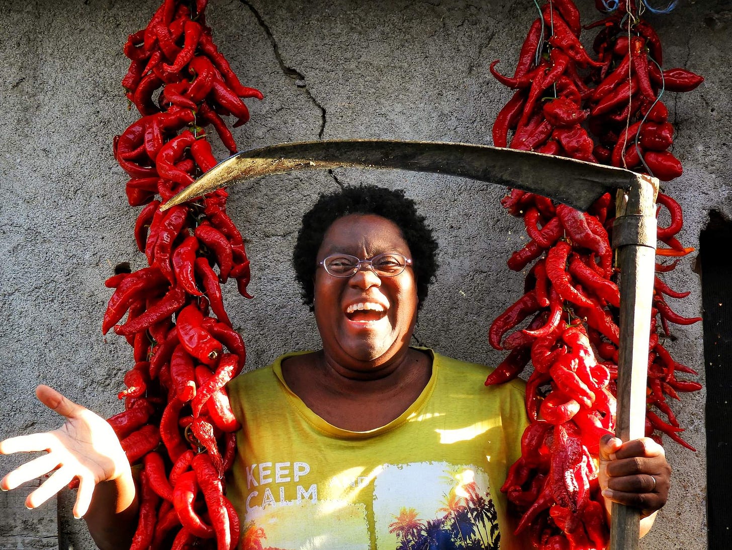A smiling Karen holding a scythe as she stands in front of a wall framed by two long strings of red peppers. 