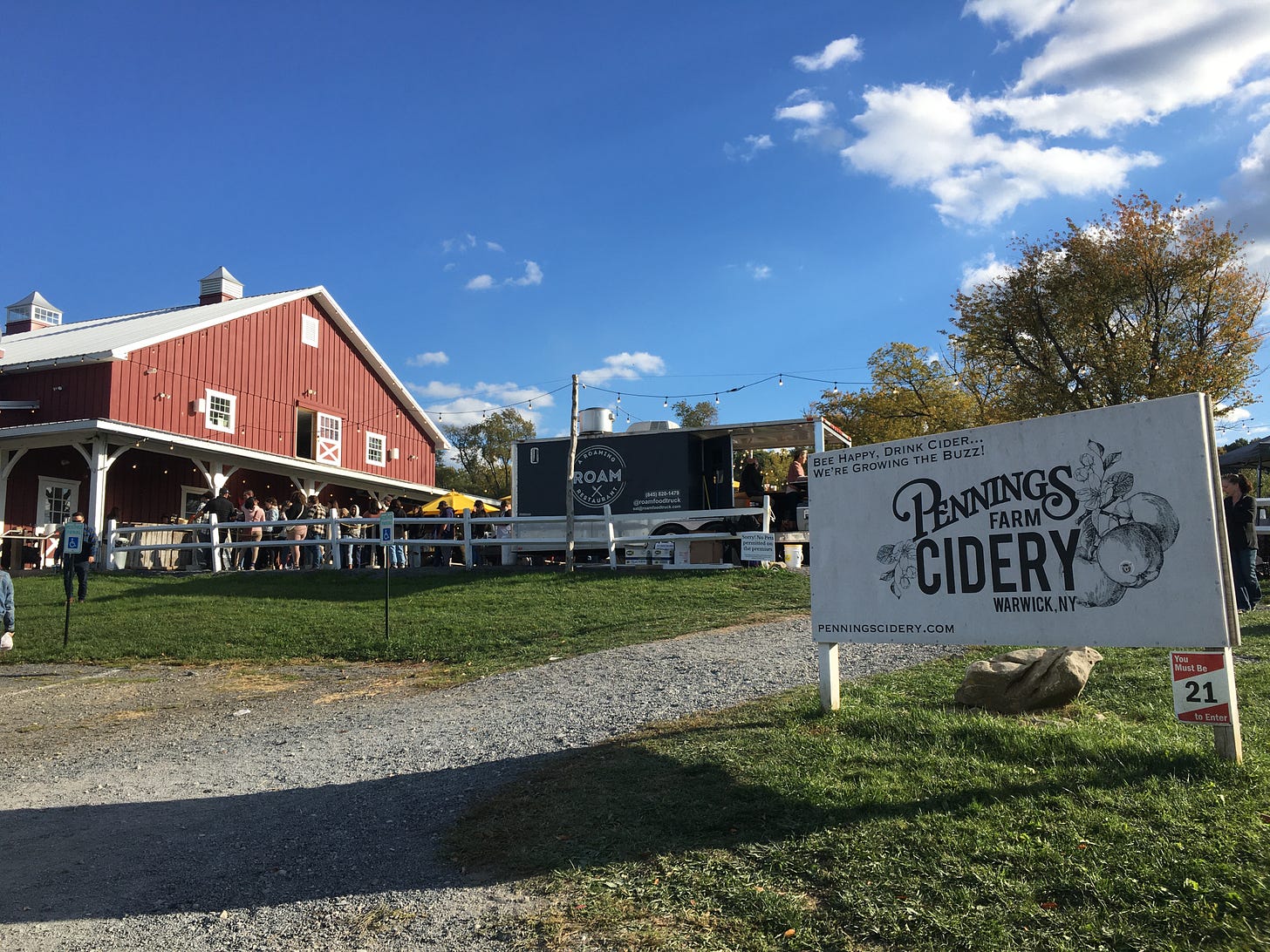 A gravel road leads to a big red barn atop a hill. The sky is blue and there are white puffy clouds in the sky. A large white sign with black letters reads "Penning Farm Cidery"