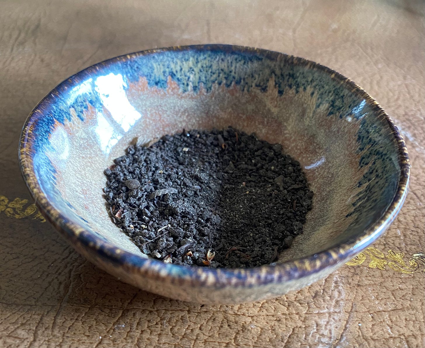 rustic pottery dish glazed with blue and brown, small  close up shot of small amount of black soil in the dish, sit-in on a leather desk mat, blurred in the background