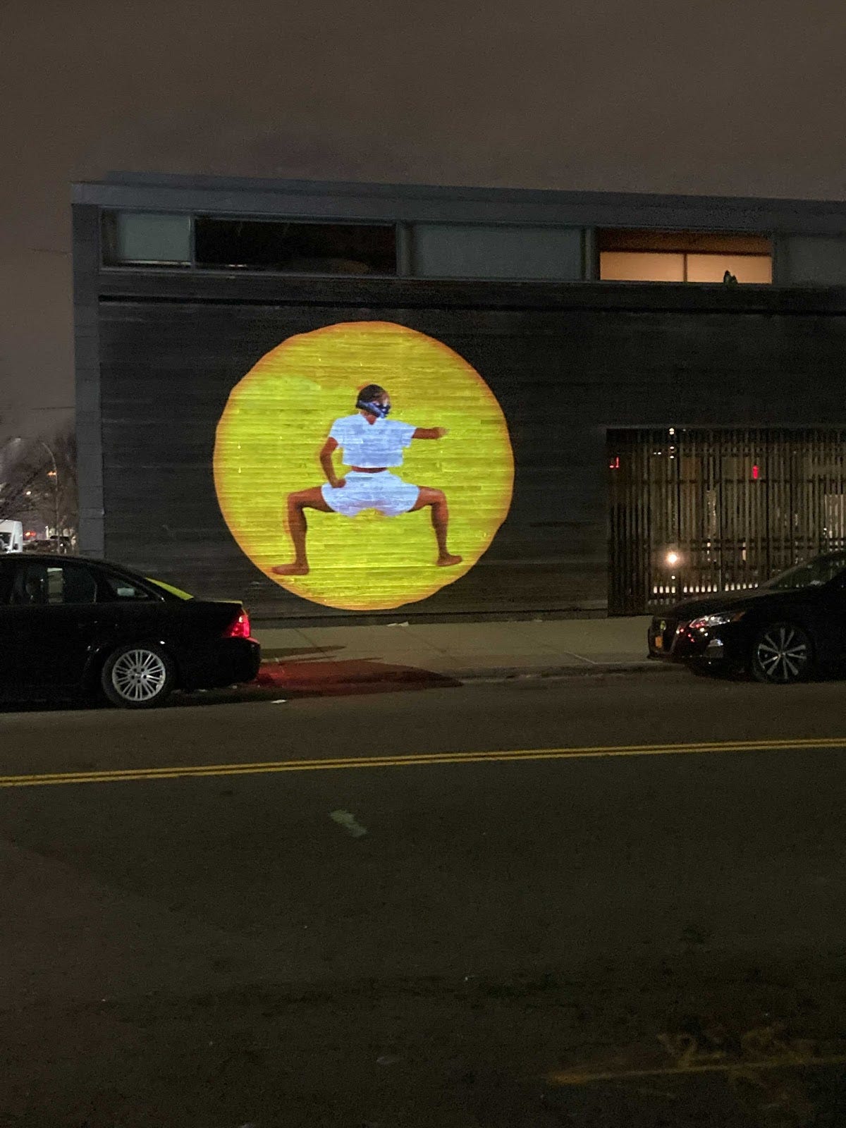 Image description: This is a projected image of a Black woman, wearing a white shirt,  white shorts, and a bandanna across her face, dancing against a yellow backdrop from the NationX portal. The projection is occurring at night against a wall in a Brooklyn neighborhood.  