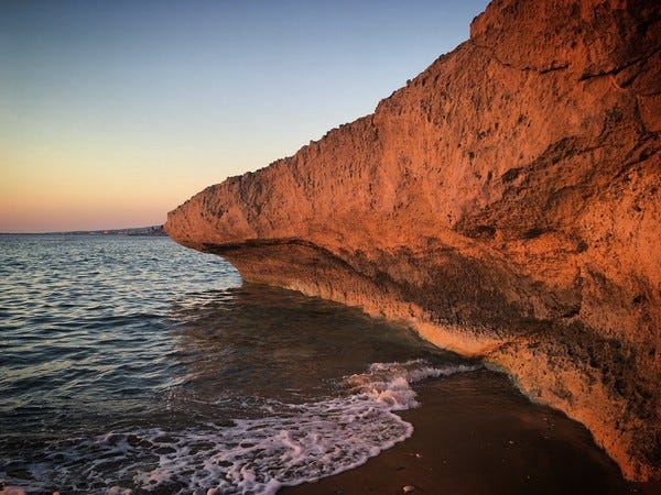 Rochedos no litoral de Pafos, no Chipre. Foto de Sergei Gussev/Flickr. Descrição de Imagem: Um litoral. O mar está calmo e o céu, sem nuvens. Um rochedo se projeta da areia com a coloração alaranjada do que parece ser o por do Sol. Ele ocupa a parte esquerda da foto e avança em direção ao meio, como uma seta.