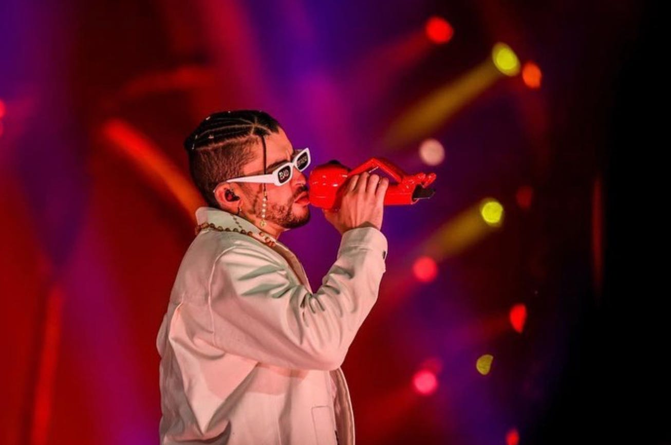 Bad Bunny at one of his concerts singing. He is wearing a beige jacket and white sunglasses. 