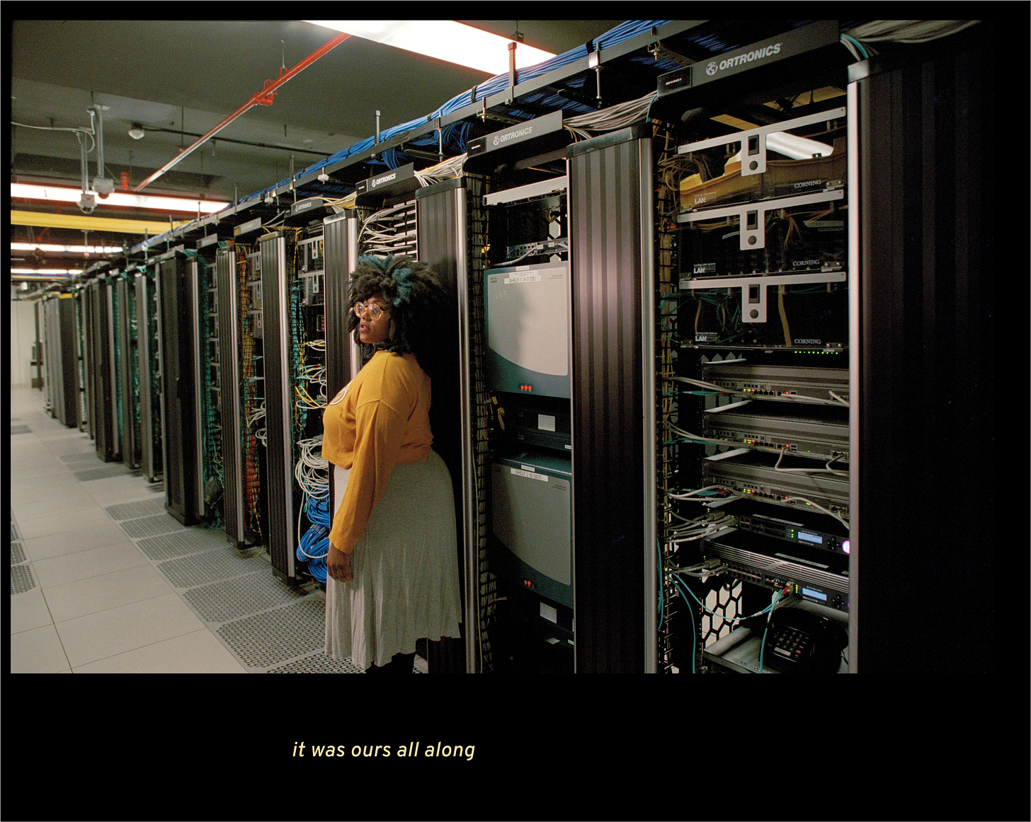 Image of a Black woman standing in a data center, in front of a wall of cables and servers. She wears a yellow shirt, big afro, and looks into the distance.