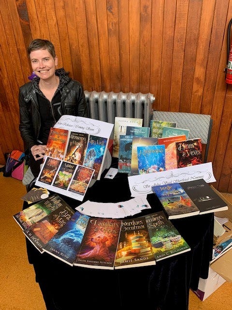 Image contains a woman with short hair and a black jacket sitting to the left of a round table covered with books. Many of the books included are the Witchy Fiction novellas.