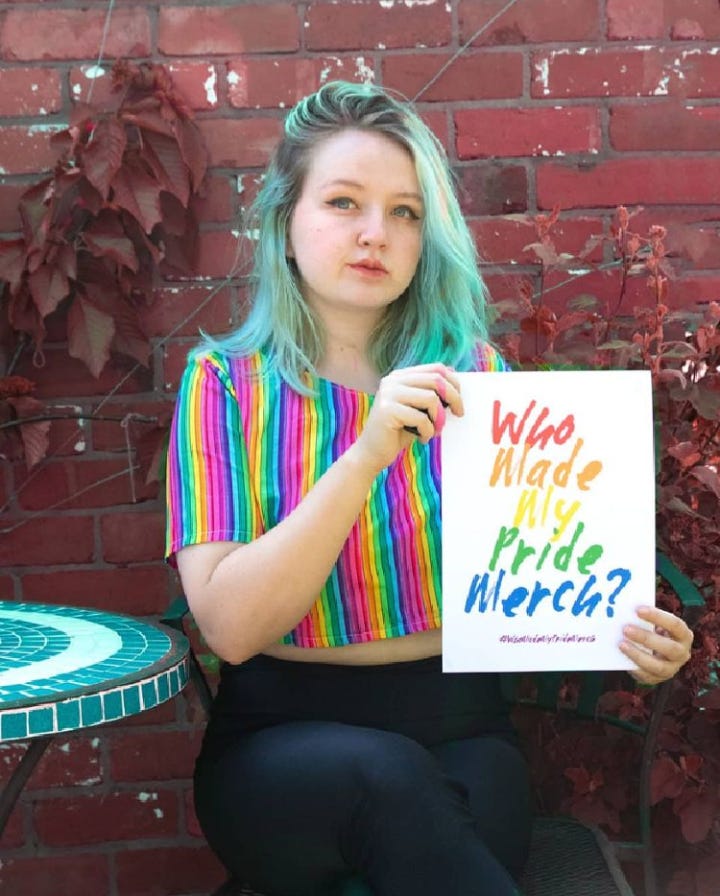 Izzy, a person with long blue hair and a rainbow shirt, sits on a garden chair holding a white poster that says 'Who made my Pride merch?' with text in rainbow colours