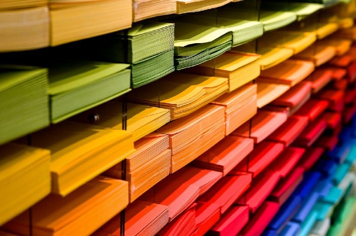 Shelves filled with drawers of slightly different colored paper. The top row is sheets of brown paper in different hues, the row below is green paper in different hues, then yellow, orange, pink, red, and blue.