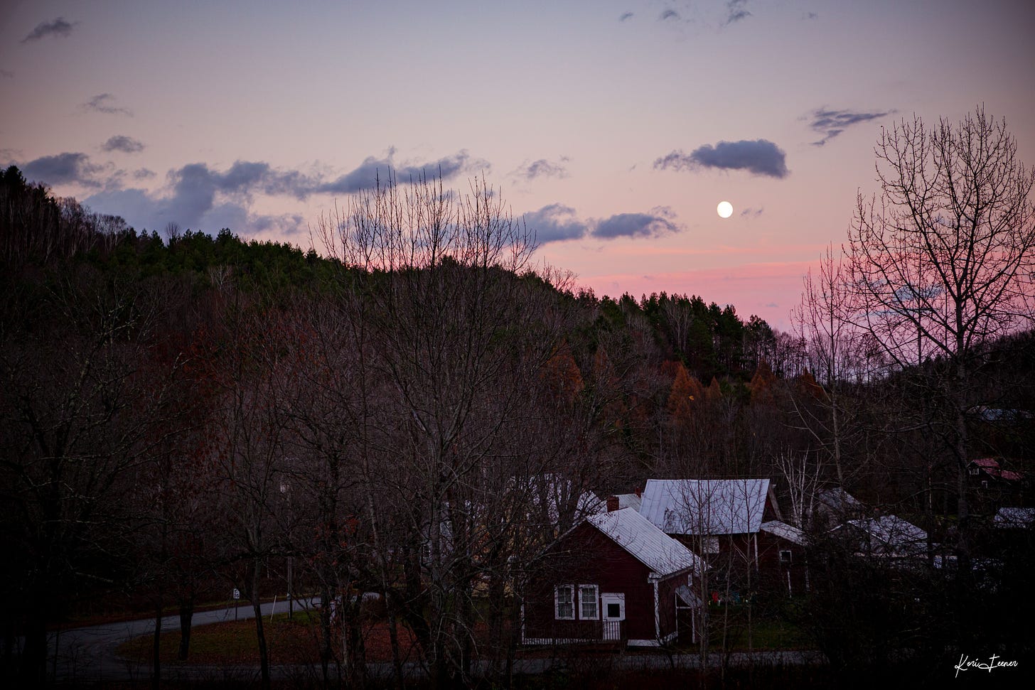 Moonrise over a rural village in Vermont. Sky is filled with pinks and blues and the trees have lost all their leaves. A schoolhouse sits in the foreground.