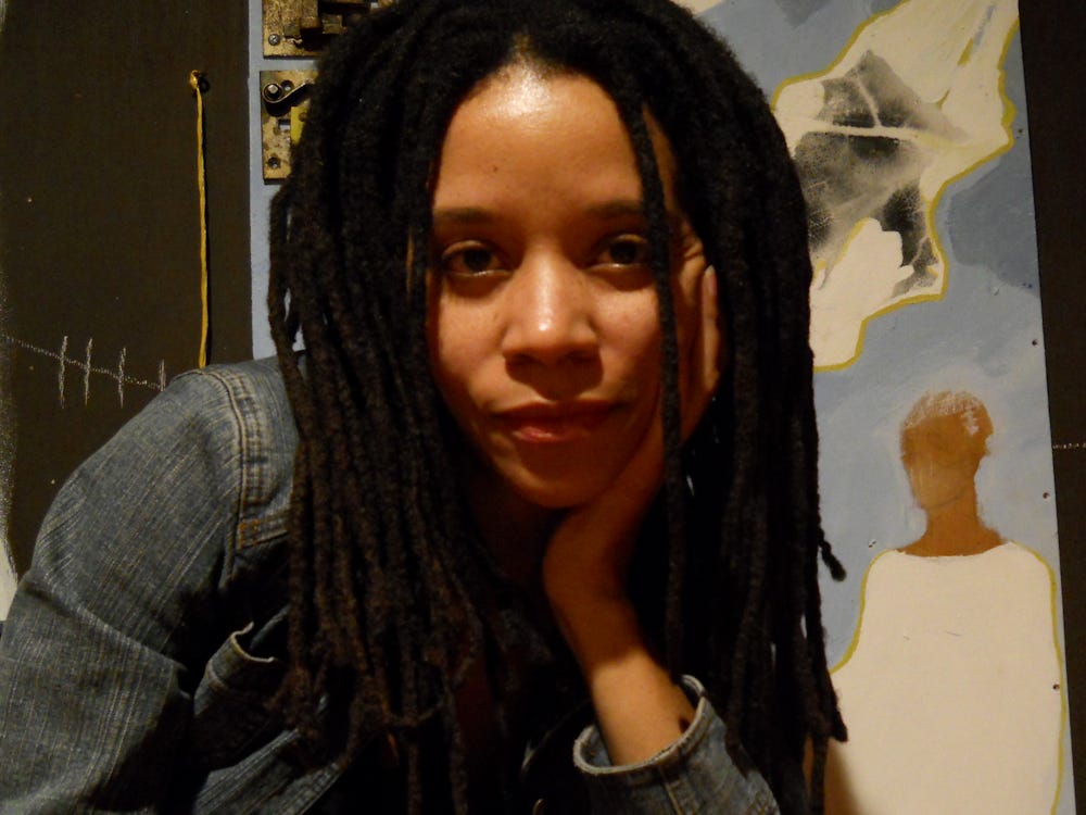 Nadïne LaFond looking directly into the camera and wearing a jean jacket set against a backdrop of one of her paintings.