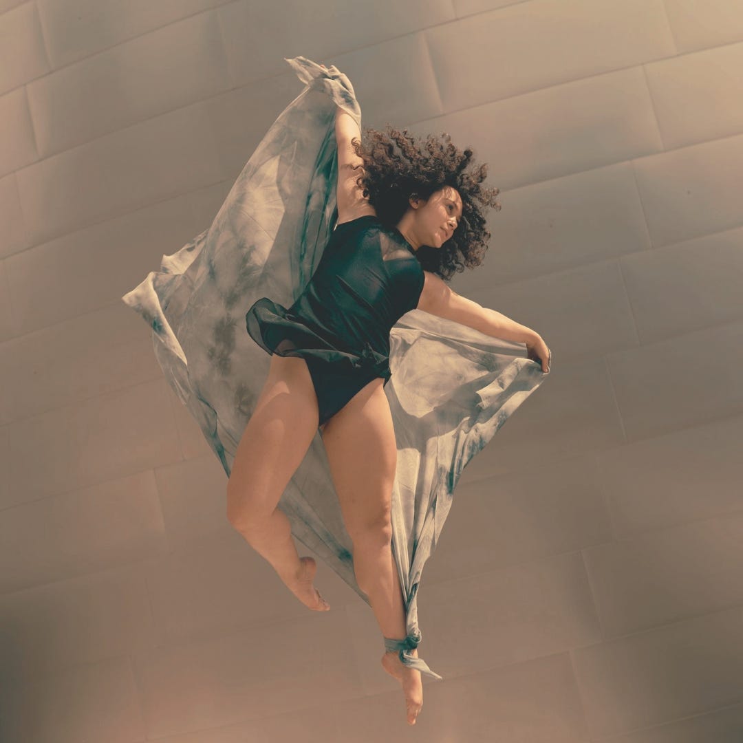  A feminine dancer is photographed mid-leap as if in flight. Her arms outstretched and her one foot touches the inside of her knee. 