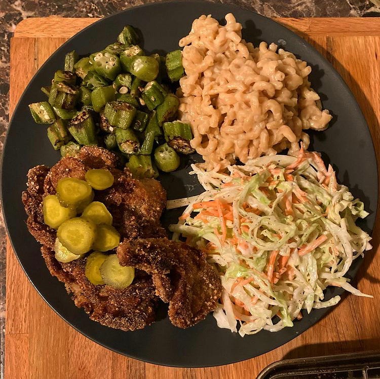 A grey plate with chicken fried oyster mushrooms, with dill pickles on the bottom left, charred okra on the top left, vegan mac and cheese on the top right, and vegan coleslaw on the bottom right