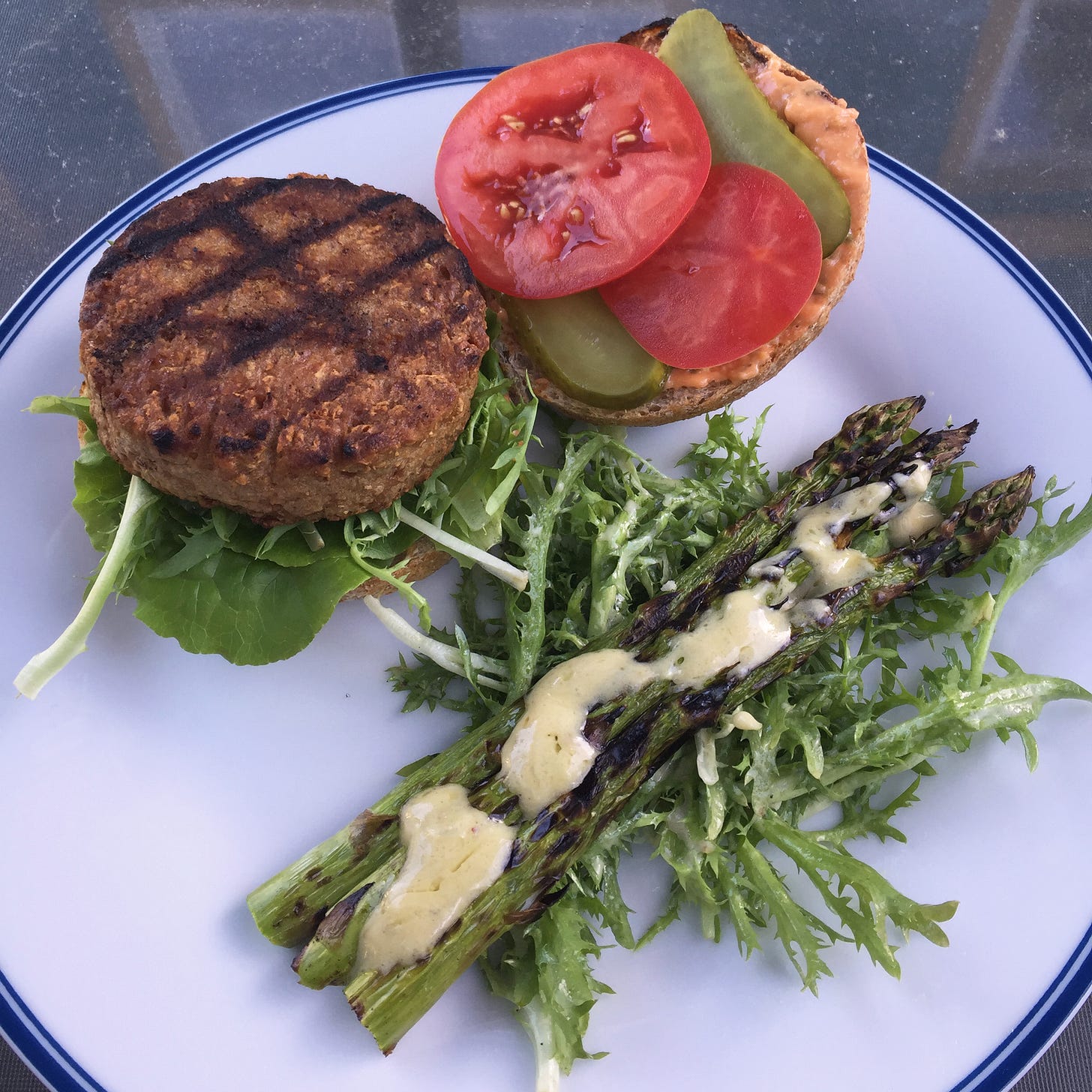 on a white plate, two halves of an open burger next to an endive salad topped with spears of grilled asparagus. The burger has a Beyond patty and lettuce on one side and orange burger sauce, tomato, and pickle on the other.
