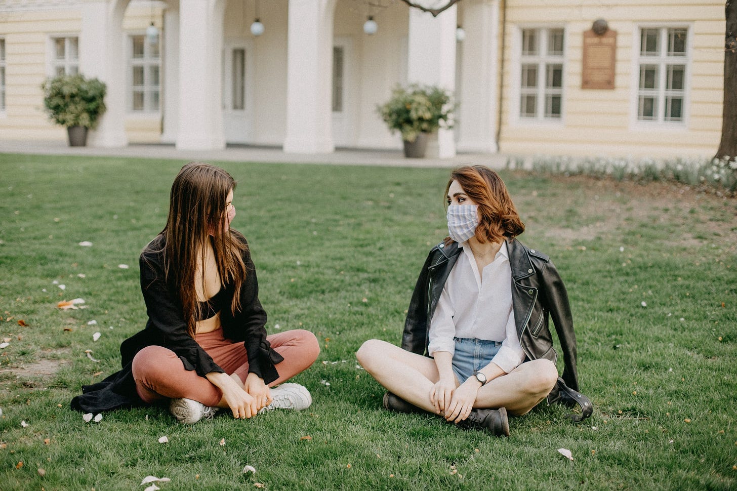 Two friends sit in a park and socialize with their masks on.