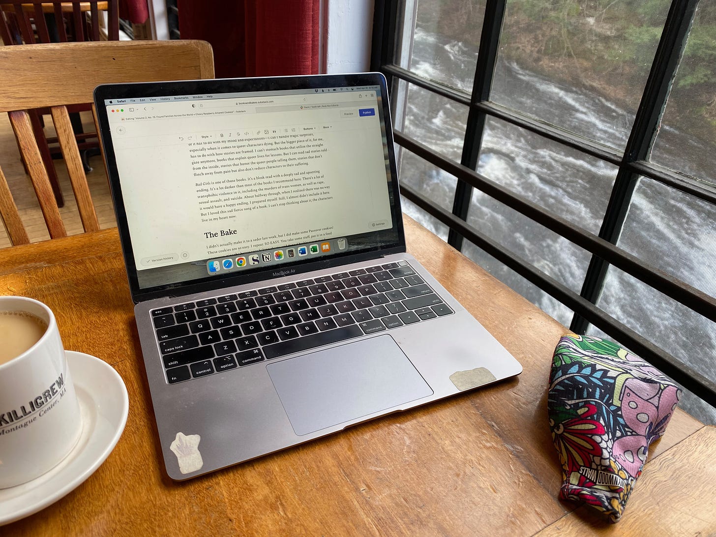 A laptop sits open a wooden cafe table next to a mug of tea and a colorful mask. Outside the window is a rushing river.