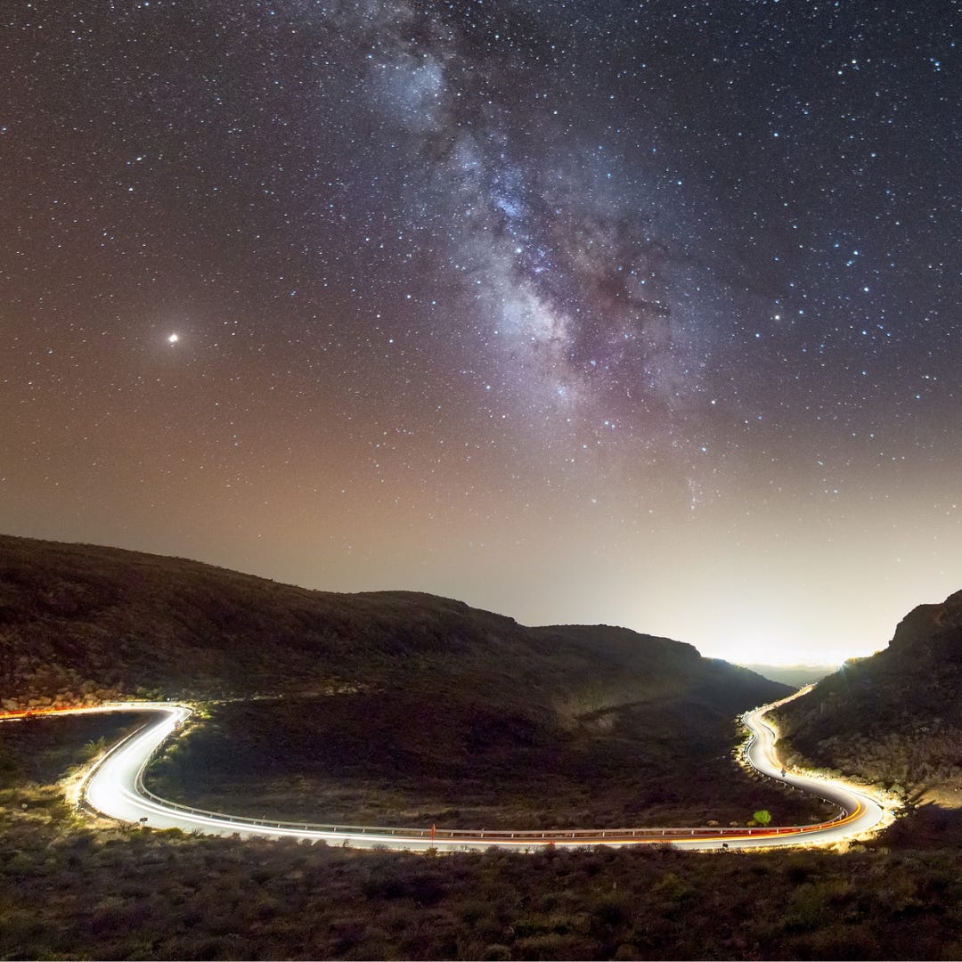 The Milky Way shines brightly over a tree-covered mountainous area. A highway runs through the mountains in the shape of a “u”. There is a golden shine to the road and the stars.The Milky Way shines brightly over a tree-covered mountainous area. A highway runs through the mountains in the shape of a “u”. There is a golden shine to the road and the stars.