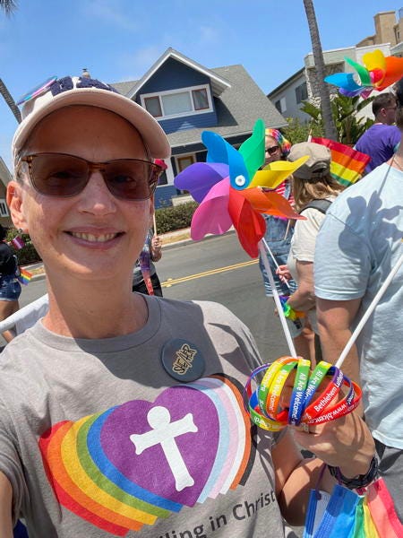 Photo of a smiling woman (me) with a t-shirt with a cross in front of a series of rainbow hearts and trans/progressive Pride flag color hearts. She is holding a rainbow pinwheel and a handful of rubber rainbow bracelets.