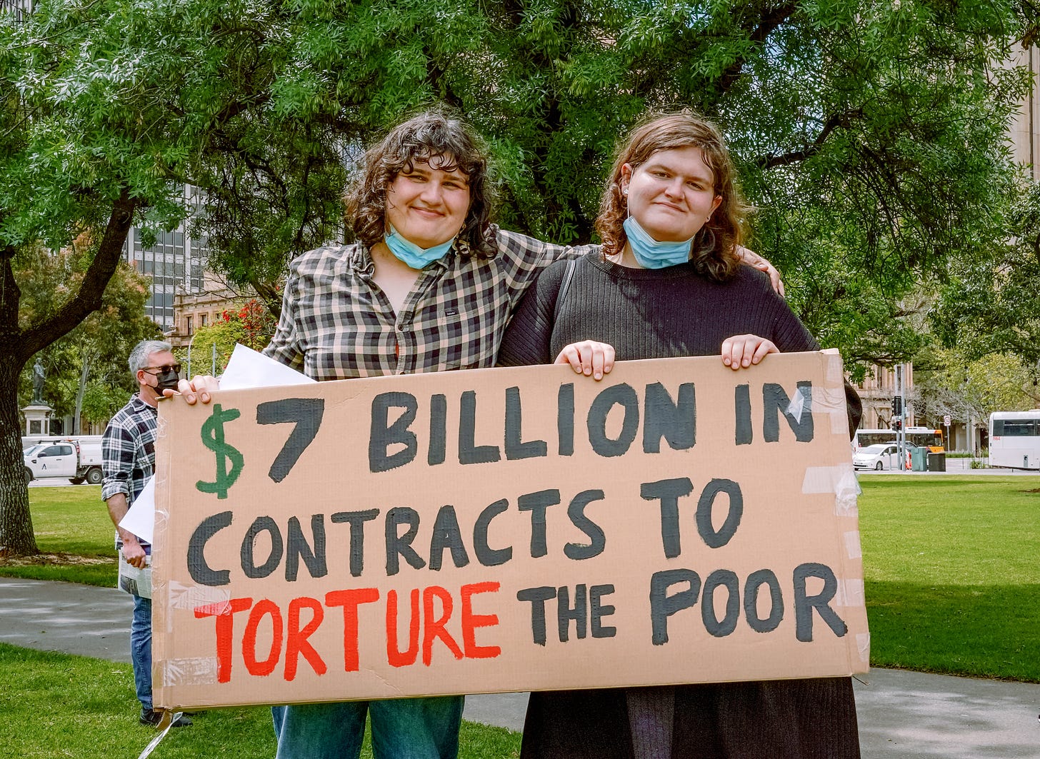 Two people holding a sign in front of a tree. The sign says "$7 billion in contracts to torture the poor"