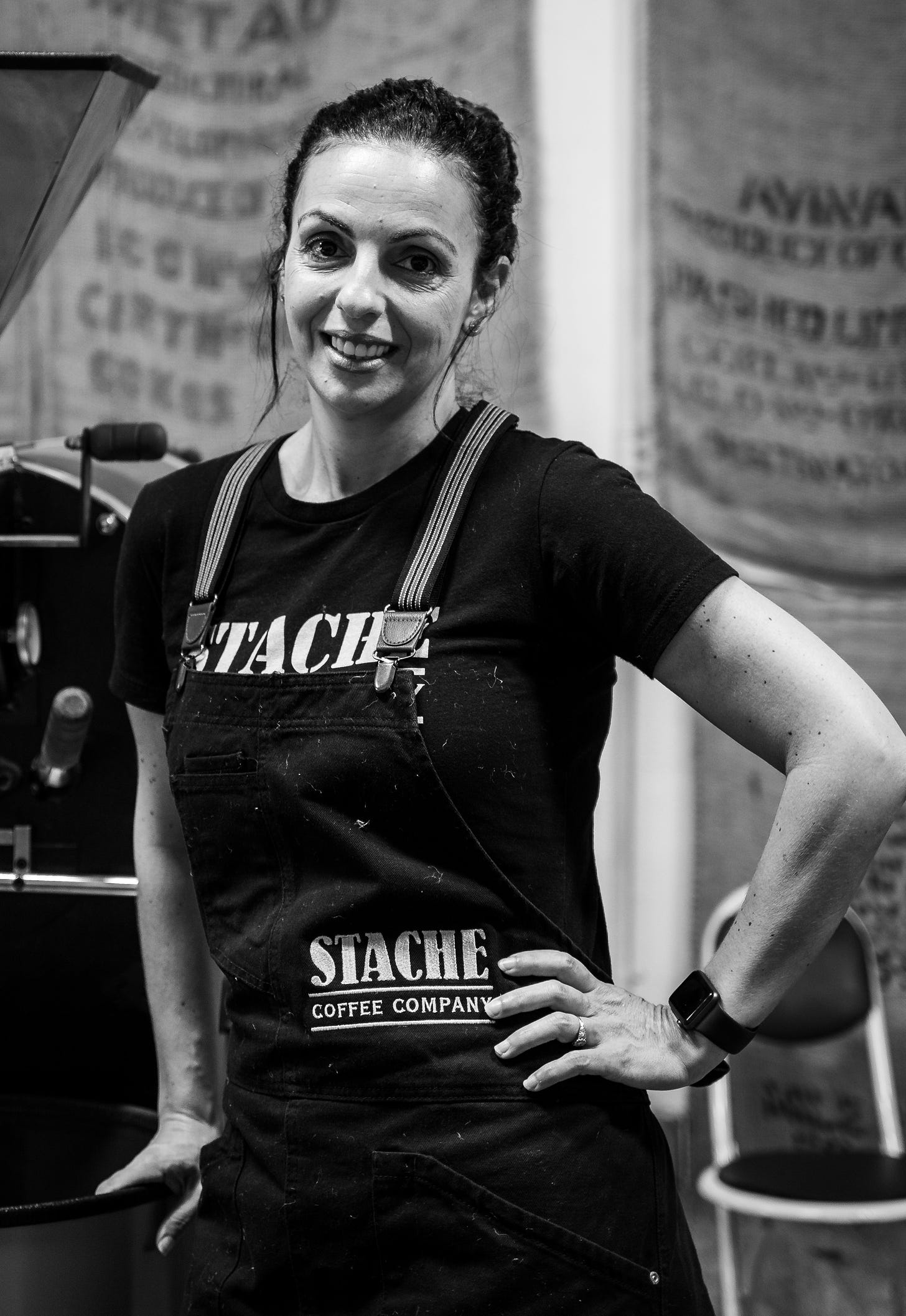 Black and white photograph of Inbal Kalin with her hand on her hip wearing a Stache Coffee Company t-shirt and apron next to a coffee roasting machine.