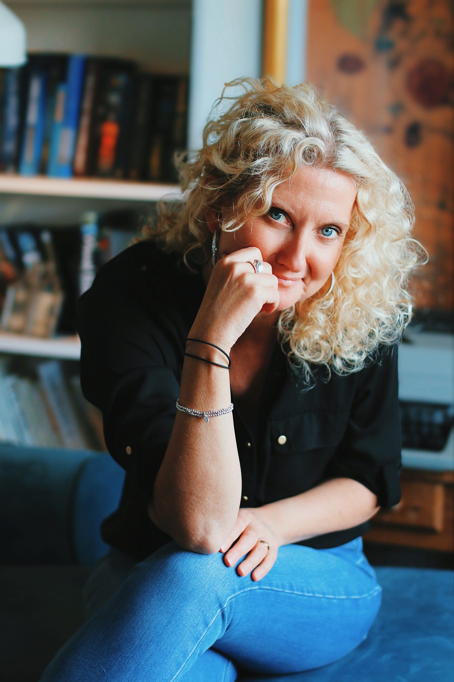 Zoe is wearing a black shirt and jeans, has amazing curly blonde hair, and is looking at the camera, smiling slightly.