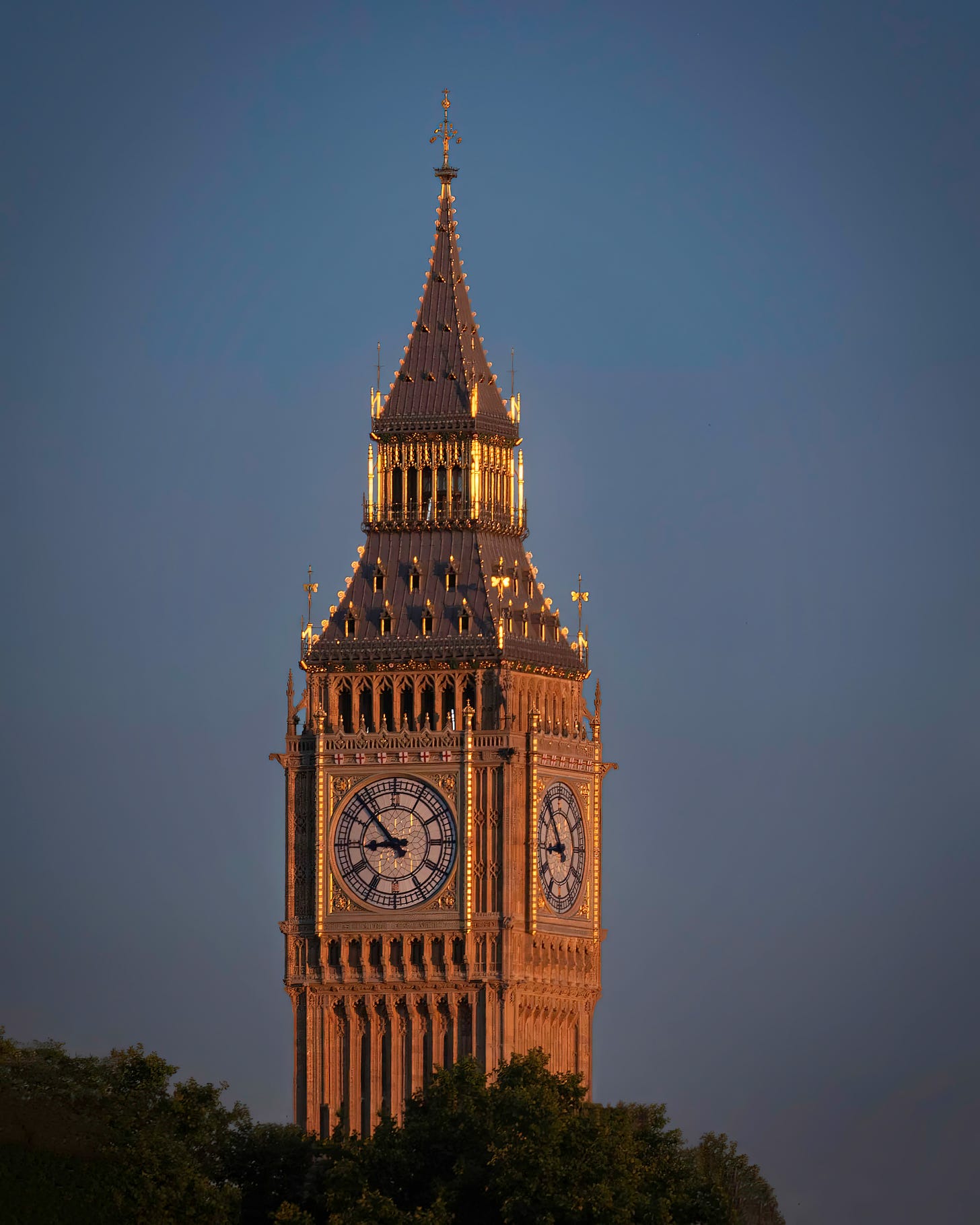 A photograph of the Big Ben on the elizabeth Tower in London with the gold lit from the setting sun against a dark blue sky with the tops of the trees in the lower foreground