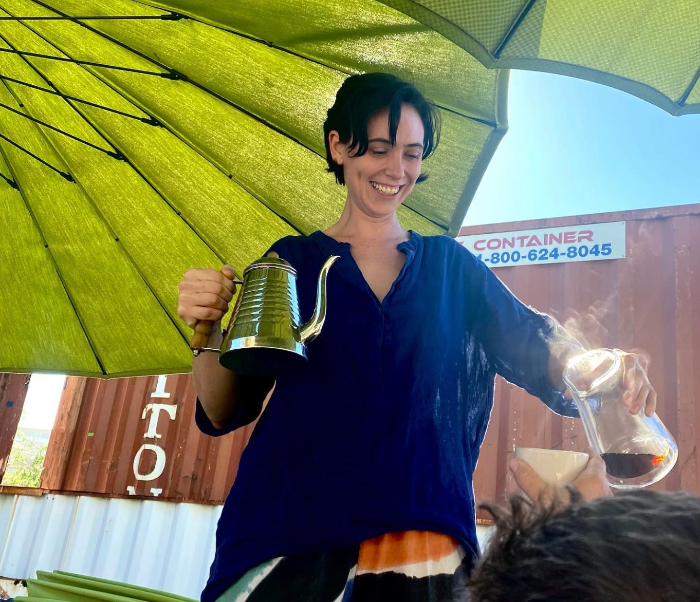 A woman with short black hair wearing a navy blue blouse under a bright green umbrella holds a stainless steel water kettle in her right hand and pours coffee out of a pot into a mug being held by an extended arm from out of frame.