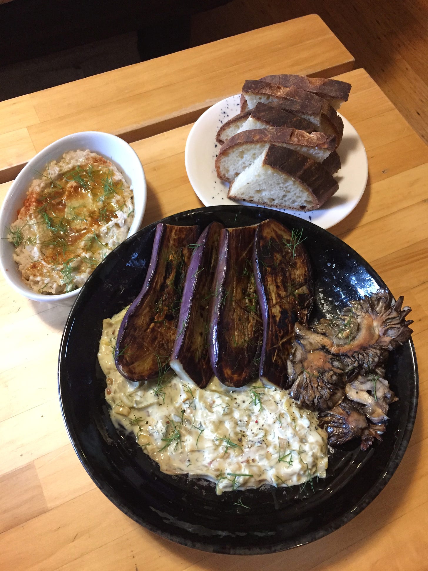 A large shallow black dish with a smear of remoulade, and nestled around it are four charred eggplant halves and big pieces of seared maitake. On a small plate are slices of sourdough with thick dark crust, and in an oblong white dish is the bean dip, coated in olive oil and dusted with spices and fennel fronds.