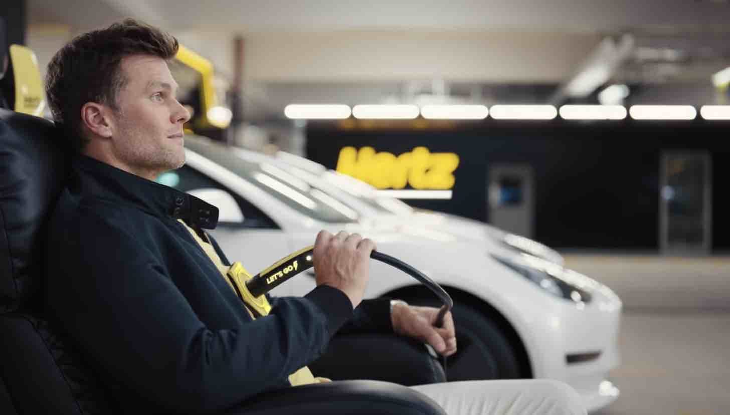 This image is a screengrab from a Hertz commercial featuring NFL quarterback Tom Brady, wherein Brady is seated amongst Tesla Model 3s in a Hertz garage. He is comically plugged into an EV charger