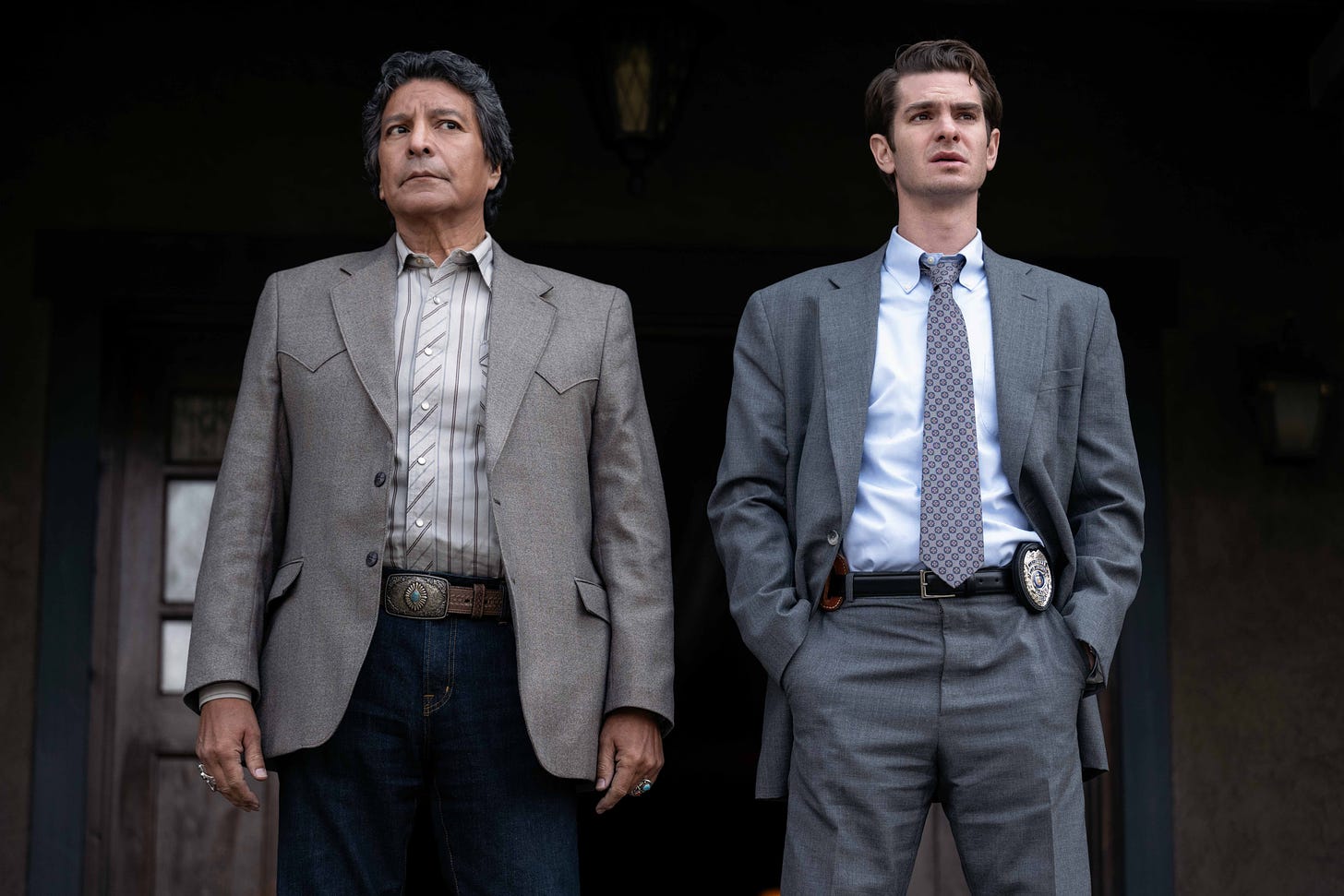 Two men stand side by side on what is apparently the front porch of a wooden house, the camera looking slightly up at them. On the left is an older Native American man dressed neatly in a Western-style sport coat, fancy belt and jeans. On the right stands a young white man dressed in a gray suit and tie. A badge is visible on this man's belt.