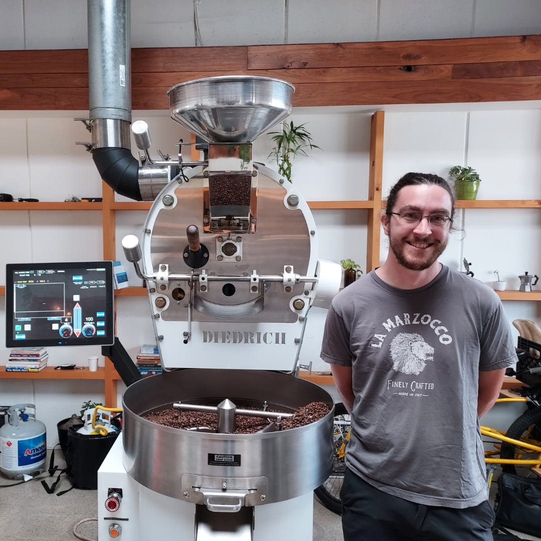 Chris O'Brien, a tall, bearded, white man with glasses and a ponytail wearing a grey t-shirt stands next to a large commercial coffee roasting machine 